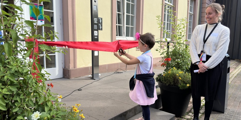 Ein kleines Kind durchschneidet ein rotes Band vor dem Haupteingang des Jugendhauses, um das Haus offiziell zu eröffnen.