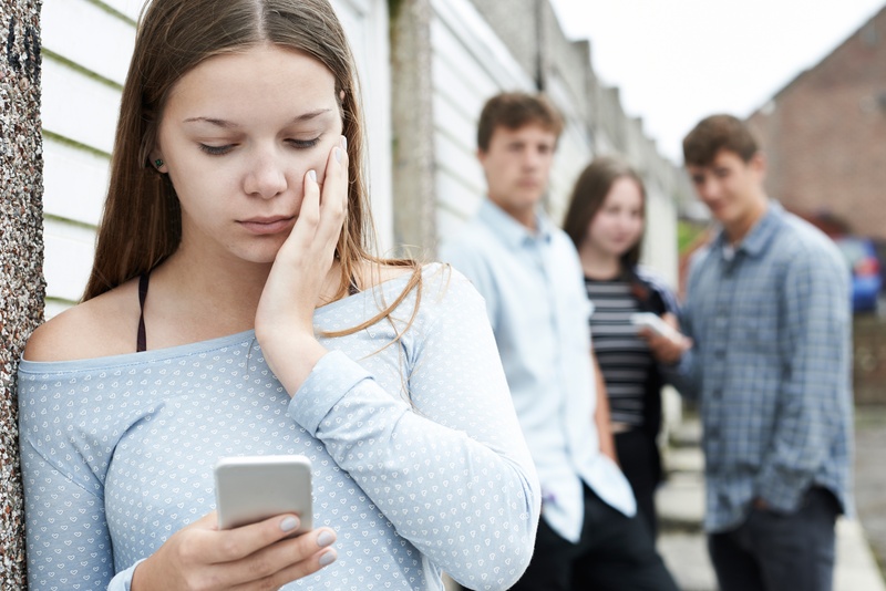 Junge Frau steht abseits einer Gruppe mit drei jungen Menschen und blickt auf ihr Handy. Es wirkt, als ob die Gruppe abfällig über die spricht.