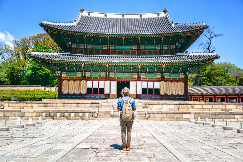 Junger Mensch steht mit Rucksack vor einem koreanischen Tempel, schaut sich diesen interessiert an.