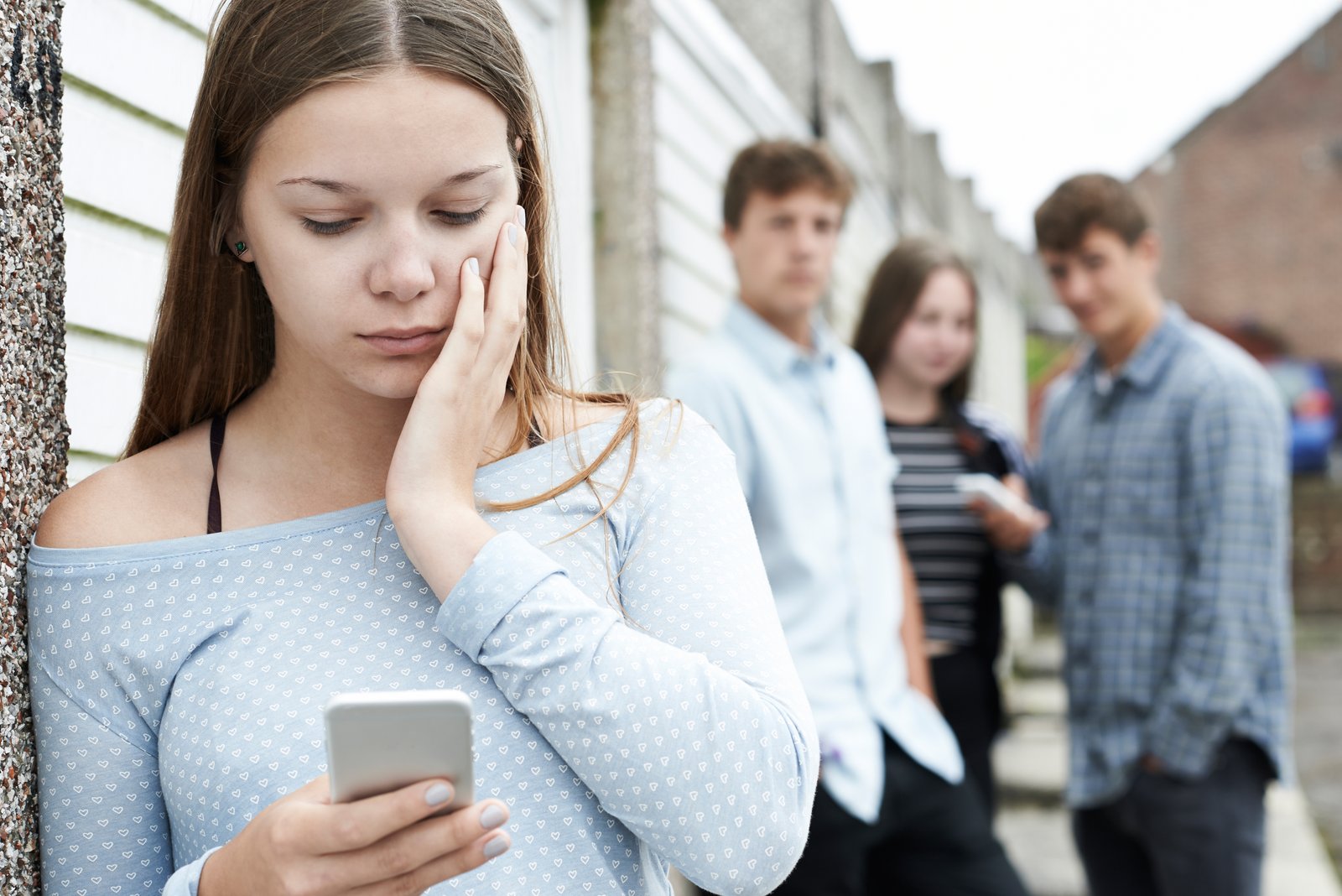Junge Frau steht abseits einer Gruppe mit drei jungen Menschen und blickt auf ihr Handy. Es wirkt, als ob die Gruppe abfällig über die spricht.
